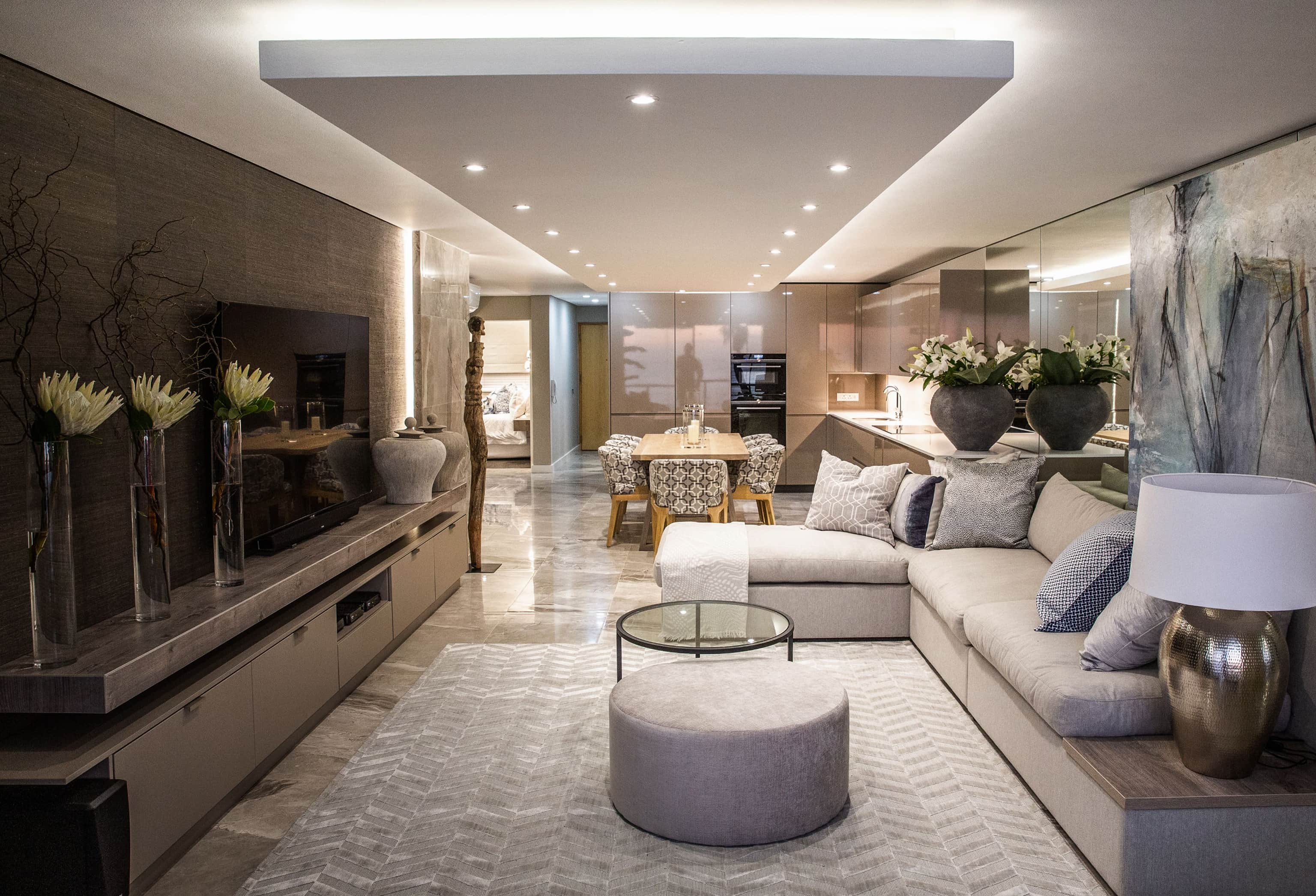 Open-plan living room with a grey corner sofa and uplit dropped ceiling, with the dining area beyond