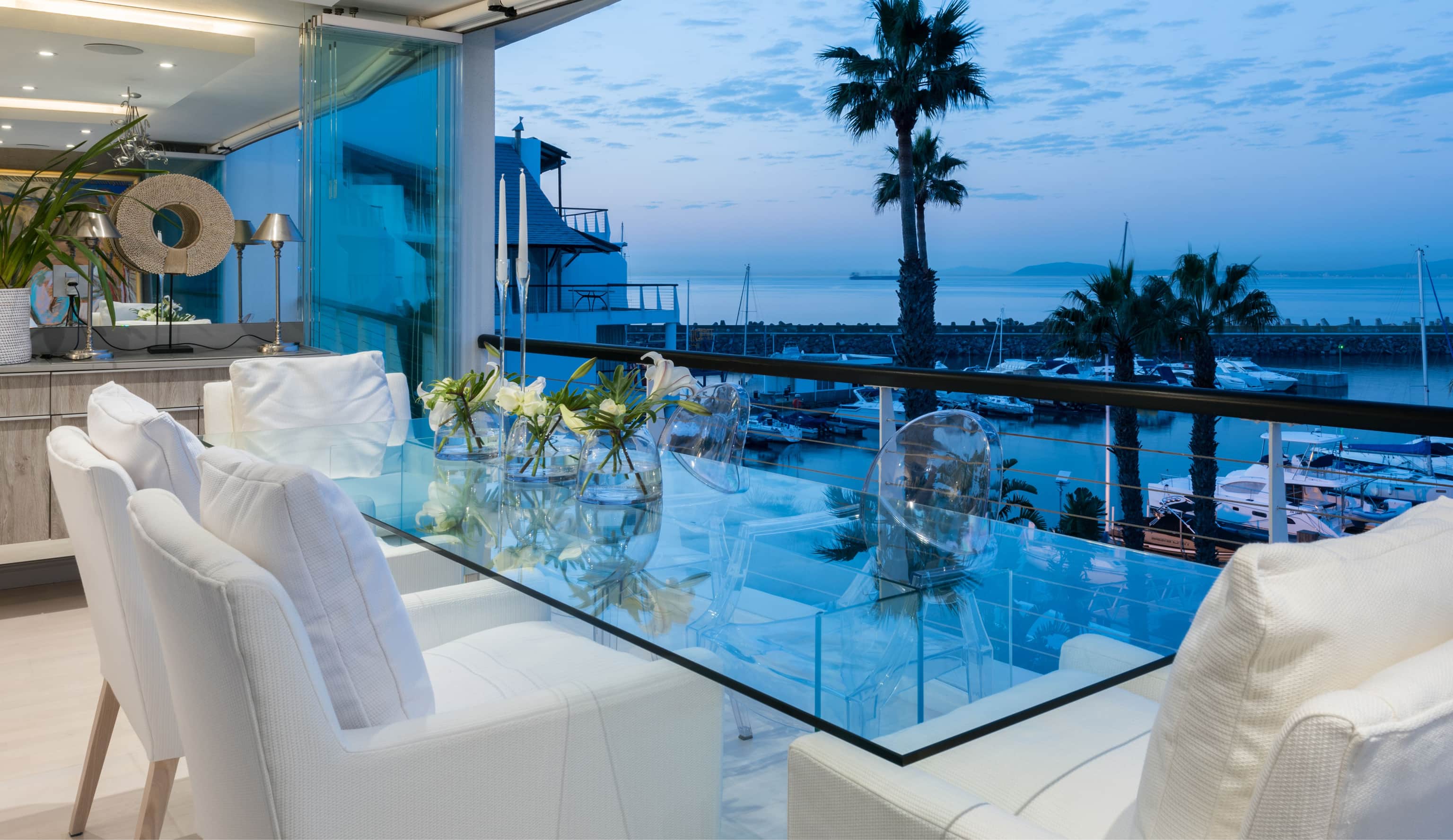Glass dining table with white chairs on a balcony overlooking the Granger Bay marina at dusk