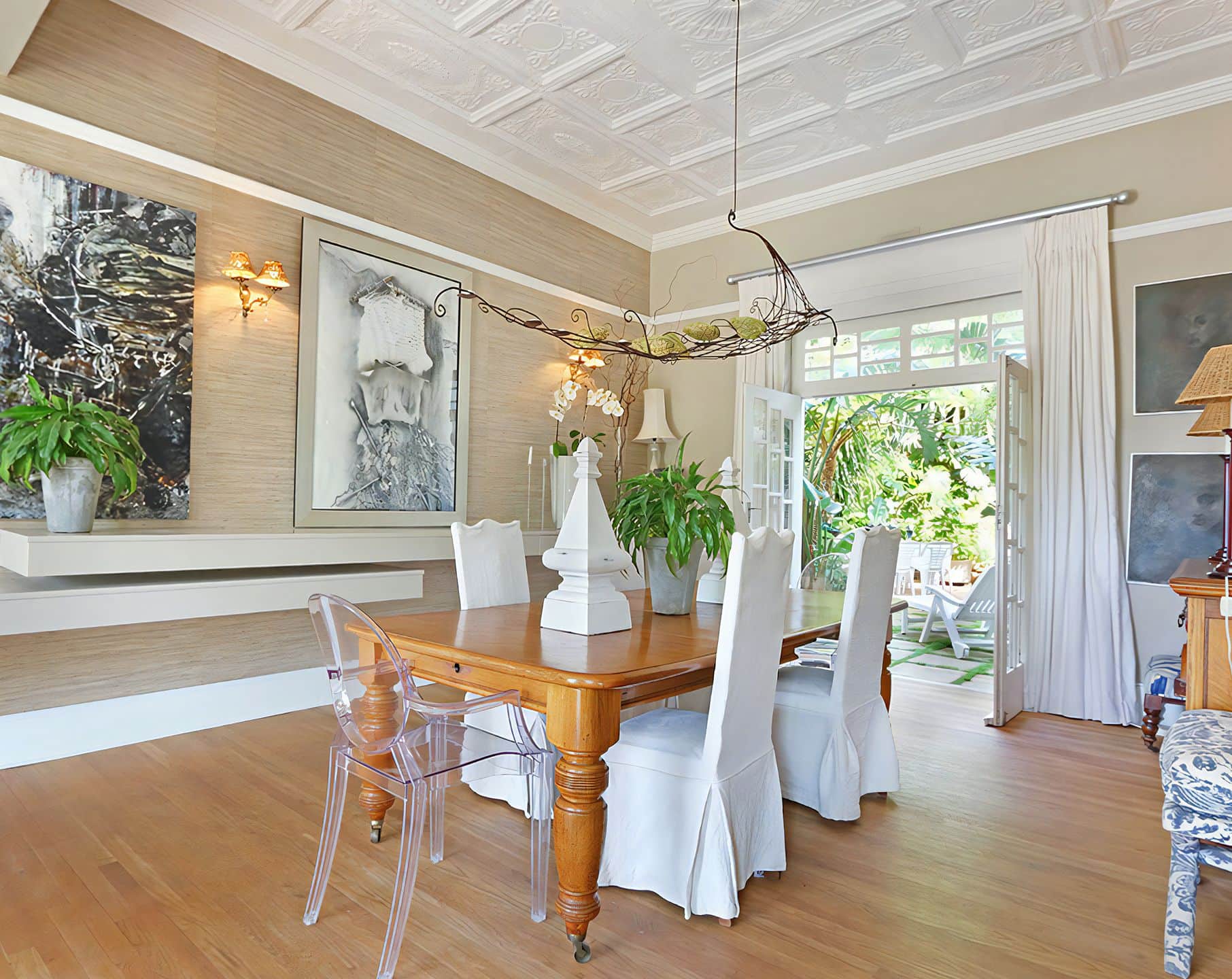 Dining room under a pressed metal ceiling, with a wooden table, white slipcovered chairs and doors open to the garden