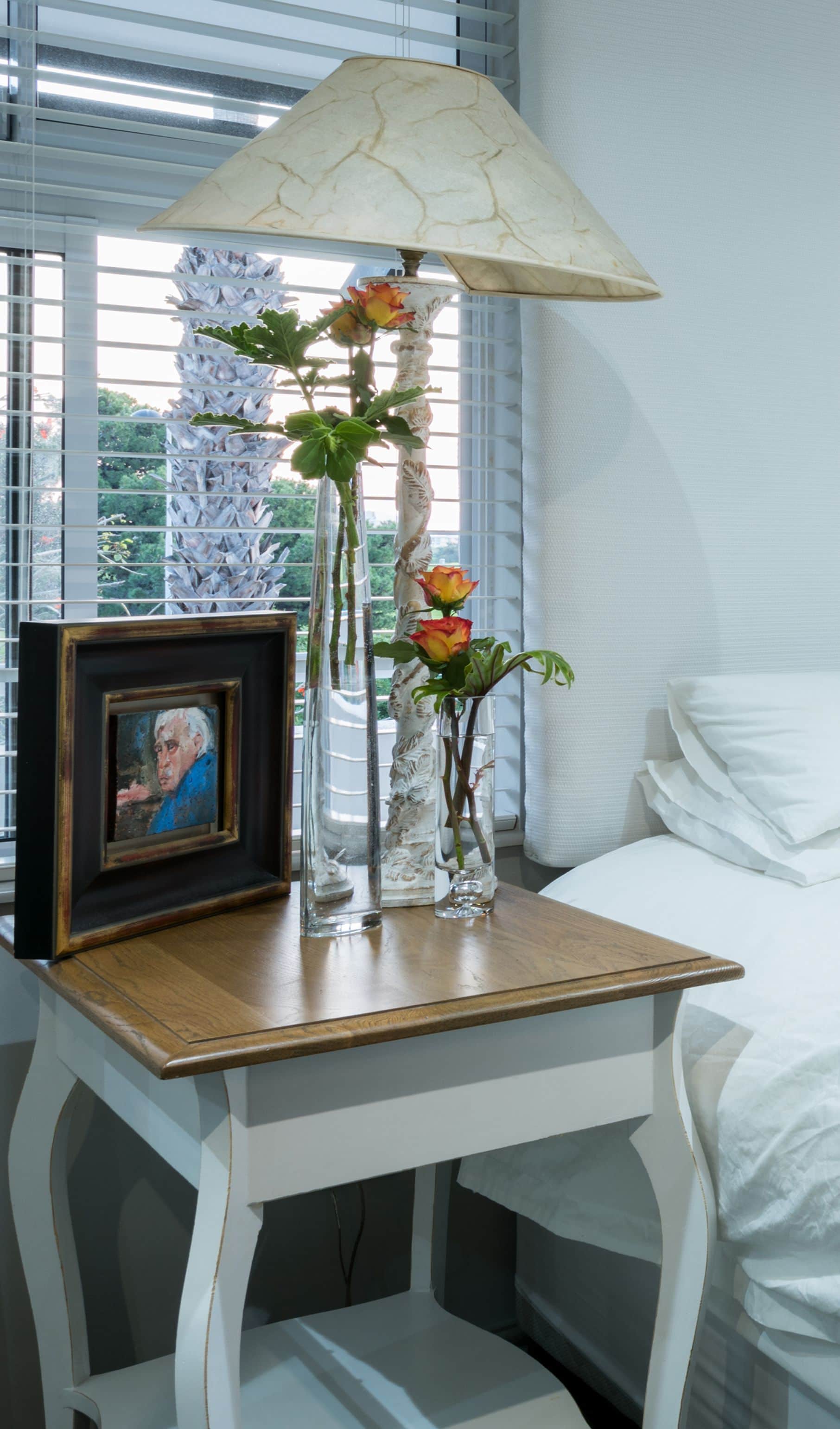 Bedside table with a framed portrait, orange roses and a lamp beside a white bed