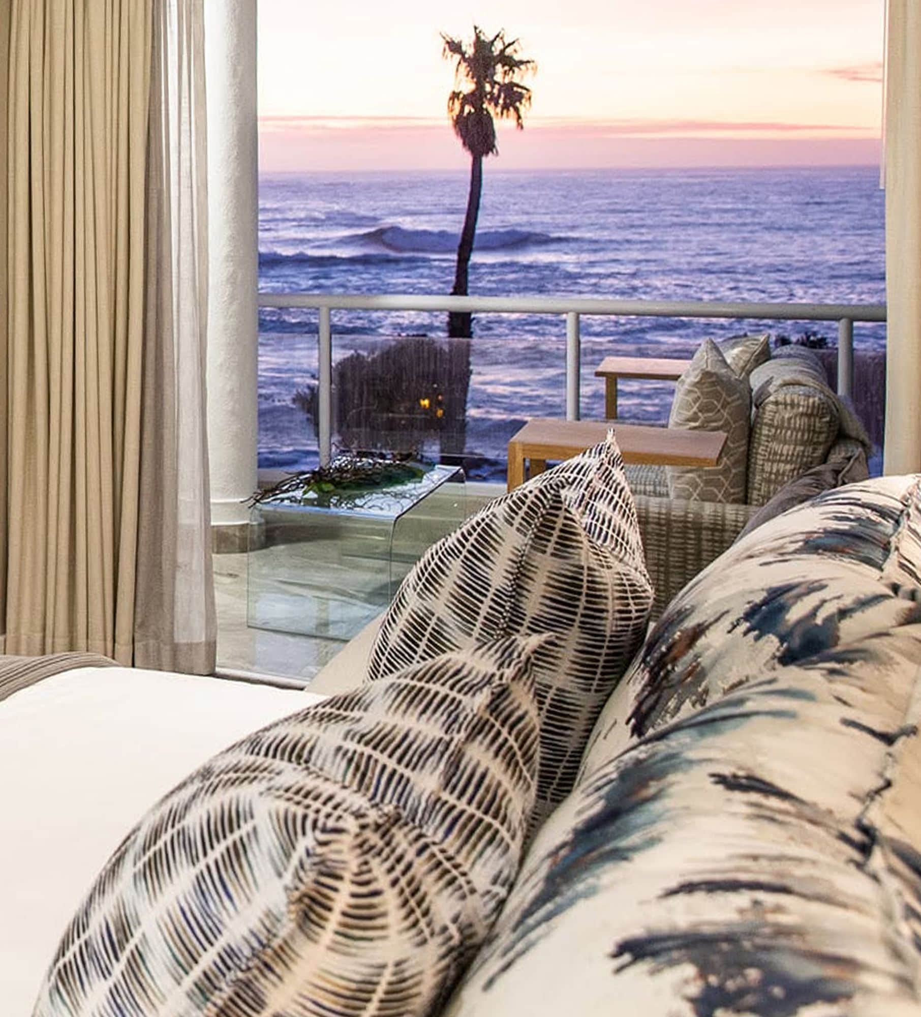 Bed with patterned cushions facing a balcony, a palm tree and the ocean at sunset