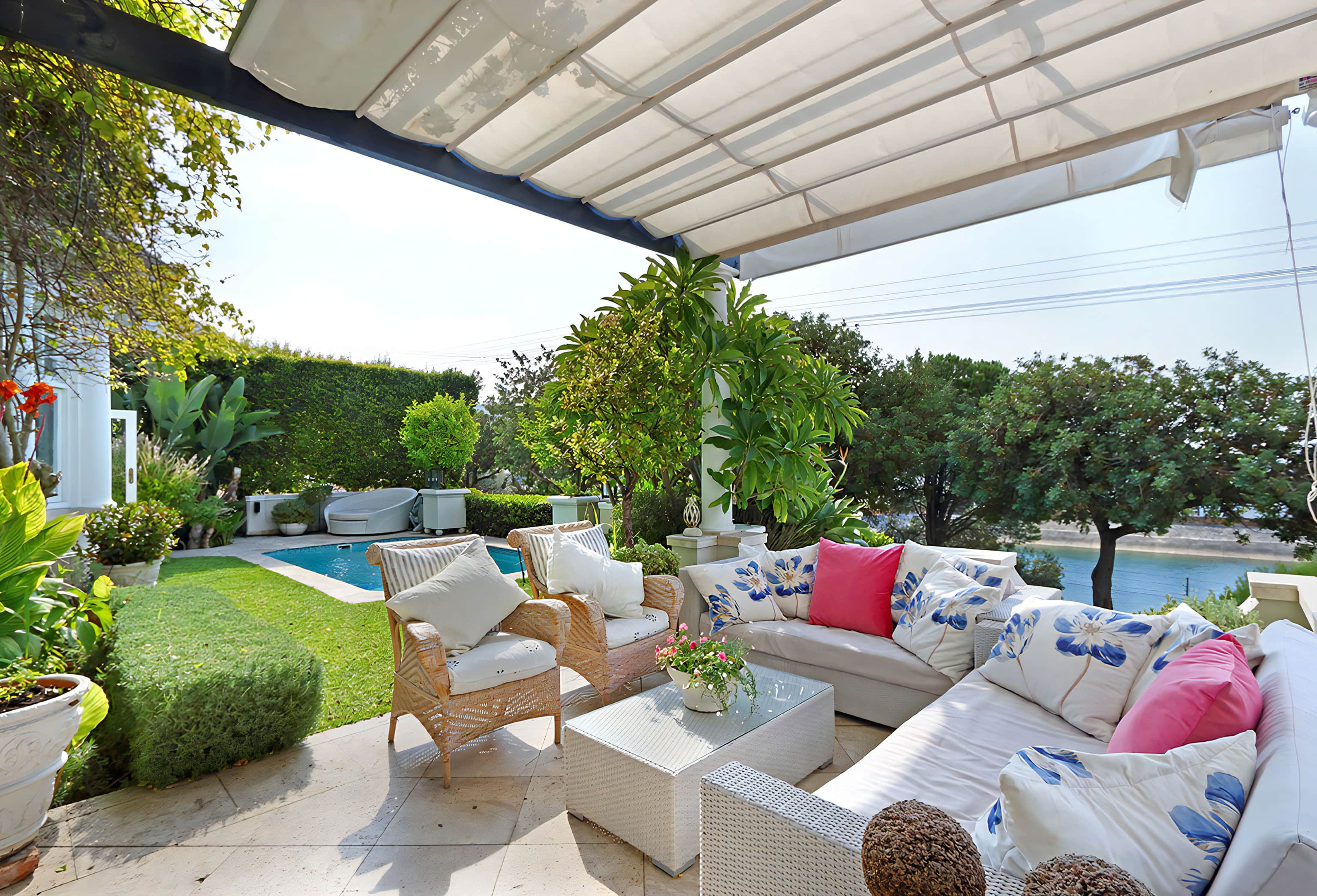 Covered patio with wicker armchairs and a white sofa with pink and blue cushions, looking onto a lawn and pool