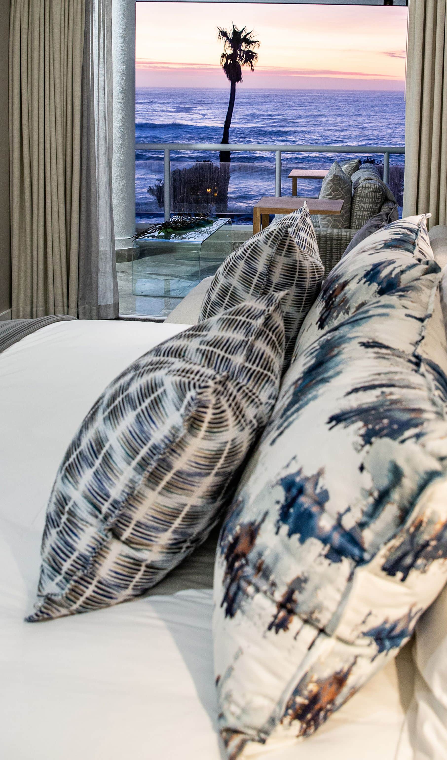Bed with patterned cushions facing a balcony and a palm tree against the ocean at sunset