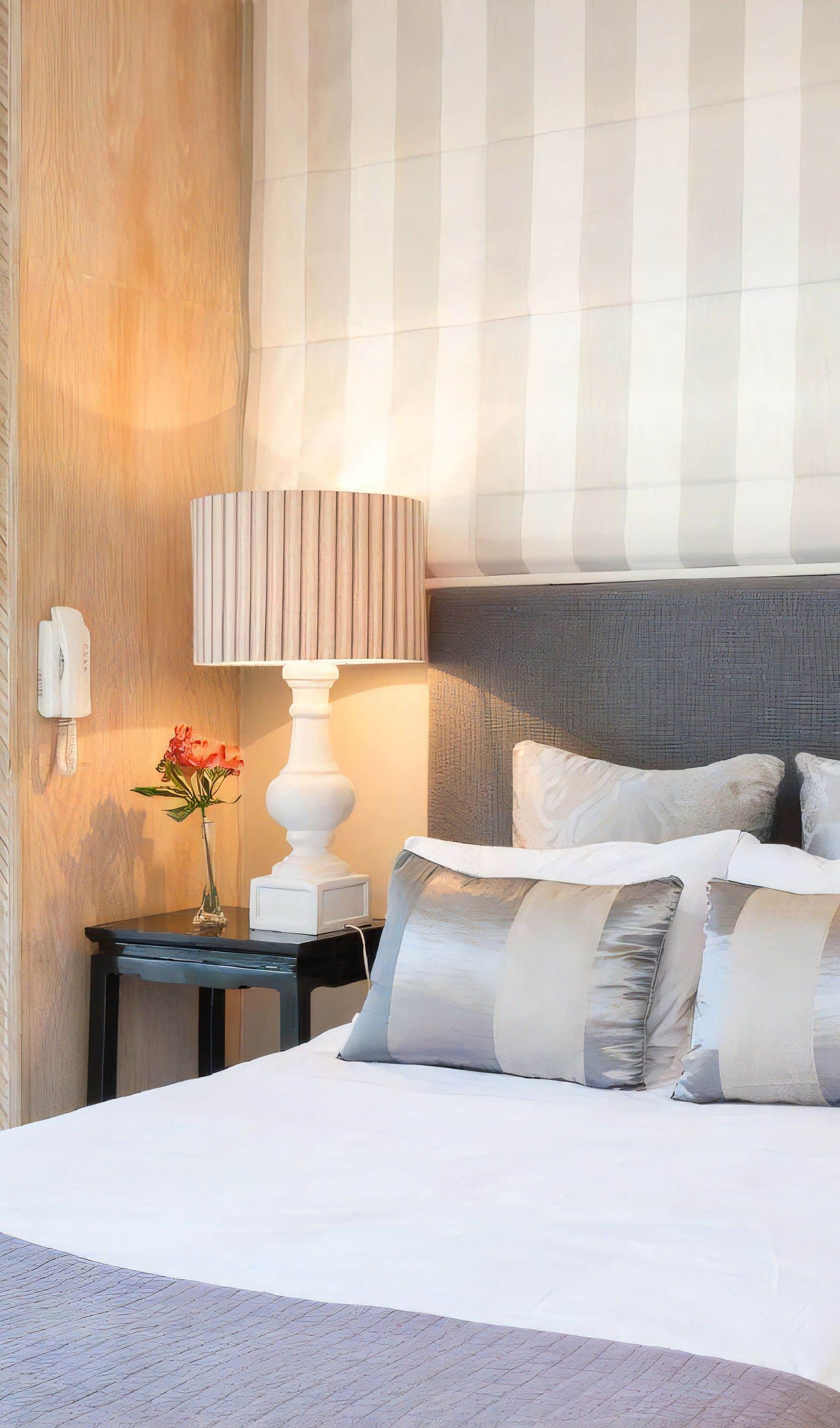 Bedside with a white turned lamp on a black table, a grey upholstered headboard and a striped blind