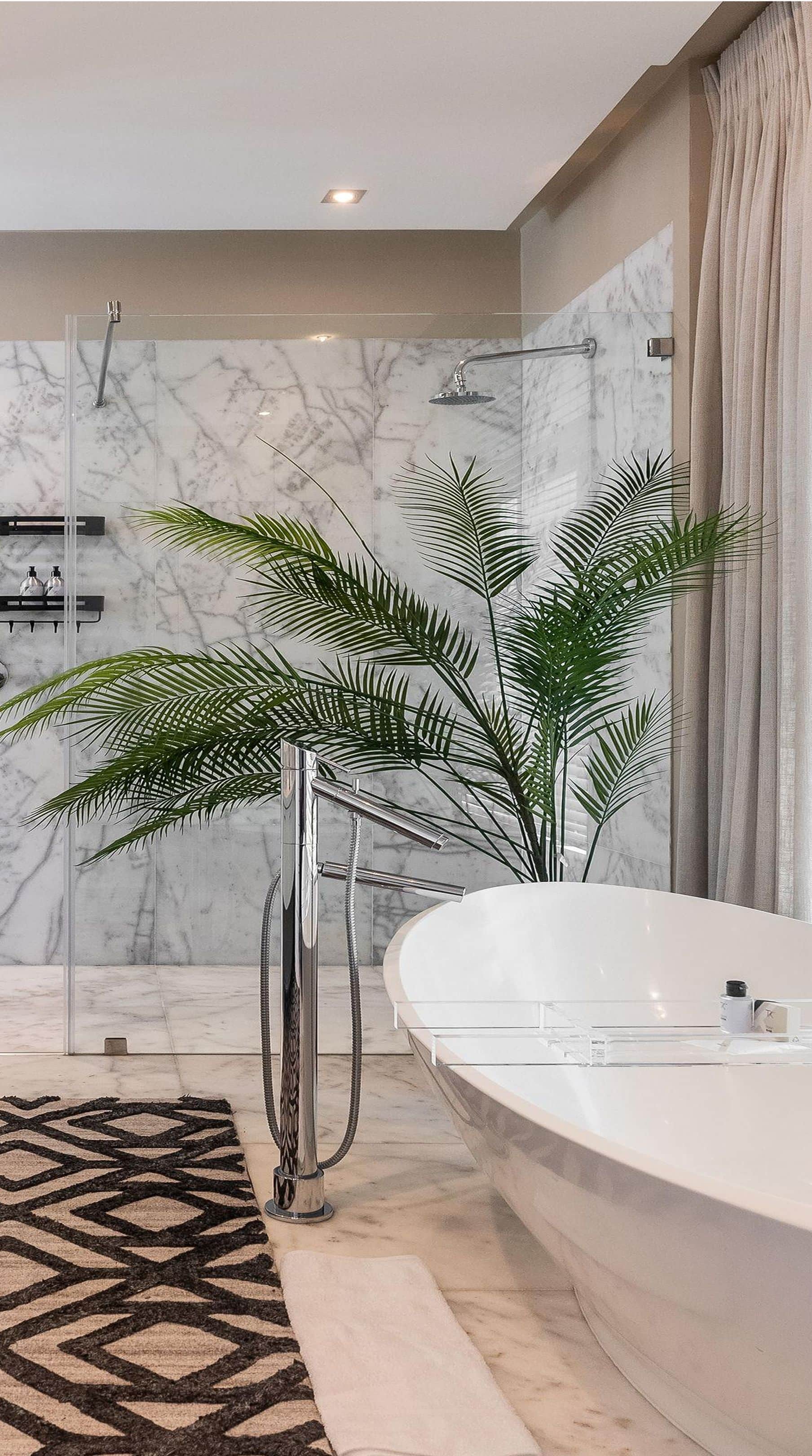Freestanding bath with a floor-mounted tap beside a palm and a marble-walled shower