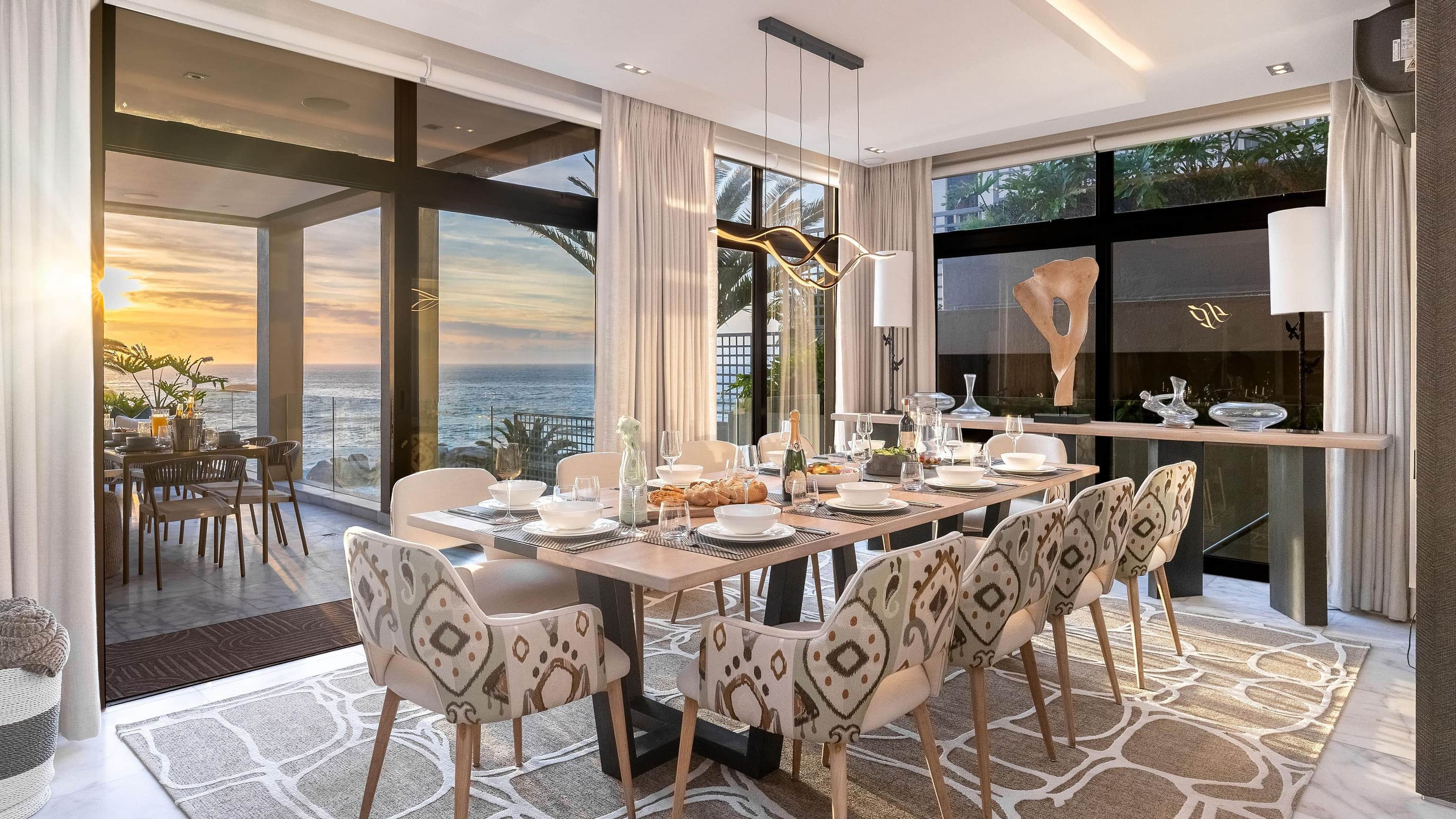 Bakoven Palms dining room at sunset, with patterned chairs around a long table and glass doors open to the ocean